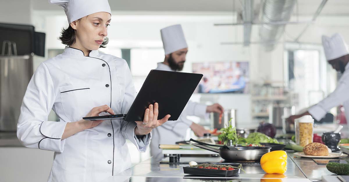 A restaurant chef holds a laptop in restaurant kitchen amongst other chefs preparing food.